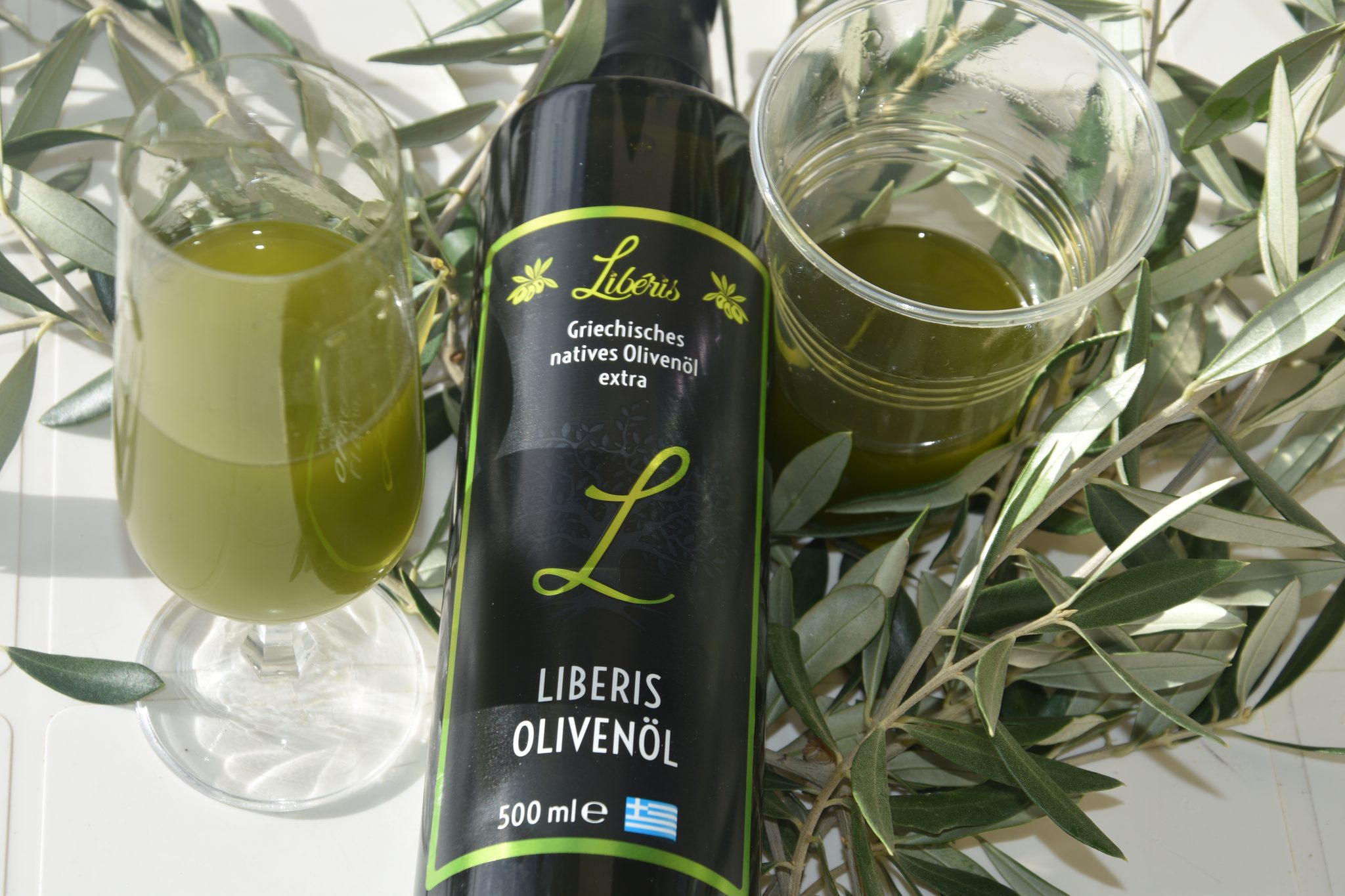 Flasche Liberis Olivenöl zwischen zwei Gläsern mit grünem Olivenöl und Olivenzweigen